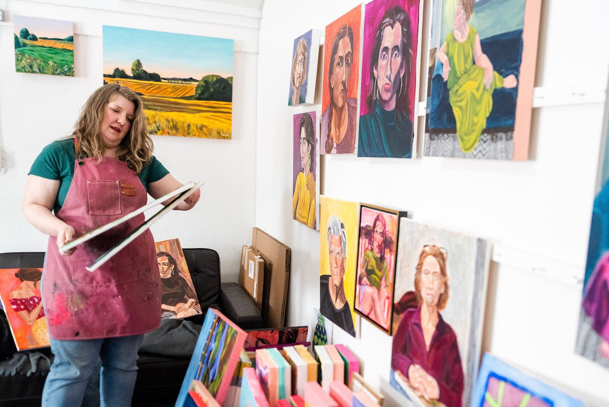 A woman wearing a pink painter's apron stands in an art studio, surrounded by numerous colorful portrait and landscape paintings displayed on the white walls.